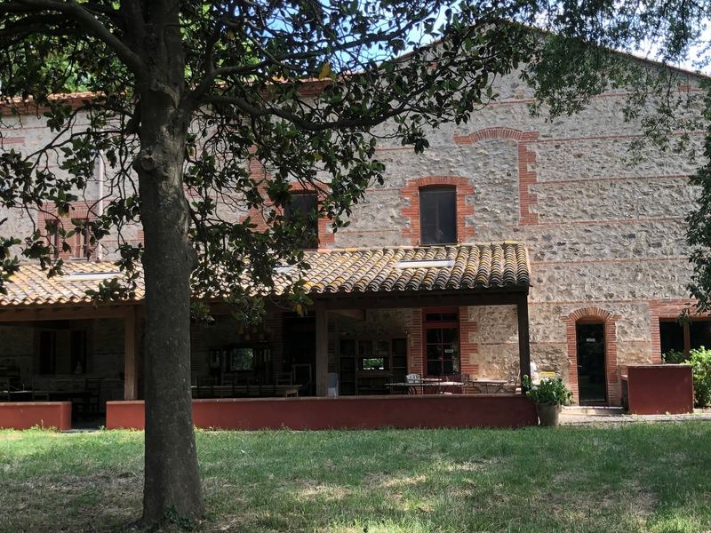 A stone and brick farmhouse with a covered, tiled terrace under a large tree.